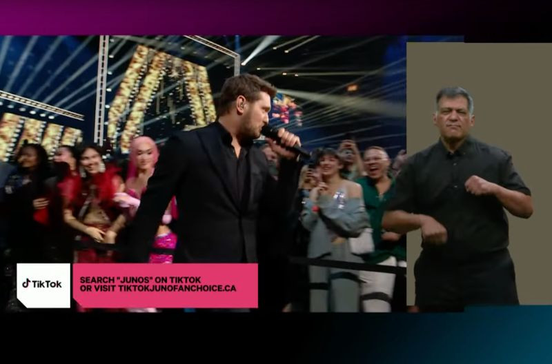 A split-screen image from the 2025 JUNO Awards. On the left, a male performer in a black suit sings into a microphone as he walks through a cheering crowd. Bright stage lights and gold-lettered “JUNOS” signage shine in the background. In the lower left corner, a pink TikTok banner reads: “SEARCH 'JUNOS' ON TIKTOK OR VISIT TIKTOKJUNOFANCHOICE.CA.” On the right side of the screen, two sign language interpreters in black clothing interpret the performance against a grey background.